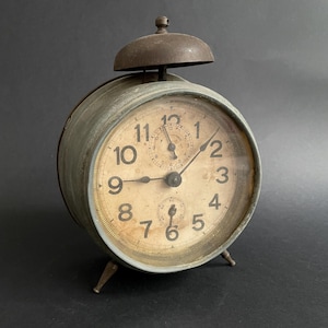 Può includere: Un orologio da sveglia vintage in metallo verde con una campana in cima. Il quadrante dell'orologio ha lancette nere e numeri arabi neri. L'orologio è su una superficie grigio scuro.