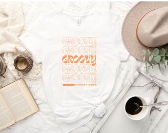 Könnte beinhalten: Weißes T-Shirt mit dem Text "Groovy", der fünfmal in einer Retro-Schrift wiederholt wird, mit den Worten "Groovy Vibes" am unteren Rand.