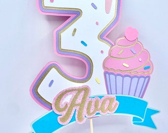 Könnte beinhalten: Ein rosa-weißer Cupcake-Tortenaufsatz mit der Zahl 3, Streuseln und dem Namen "Ava" auf einem blauen Banner.