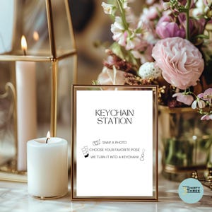 May include: A framed sign with the text "KEYCHAIN STATION" and instructions: "SNAP A PHOTO, CHOOSE YOUR FAVORITE POSE, WE TURN IT INTO A KEYCHAIN!". The sign is in a gold frame, with a candle and floral arrangement in the background.