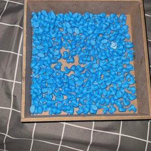 May include: A wooden tray filled with numerous small, light blue duck-shaped objects. The ducks are densely packed within the tray, creating a textured surface. The tray sits on a gray and white checkered fabric.