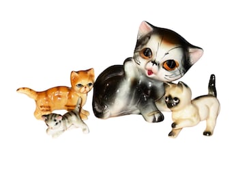 May include: A set of four ceramic cat figurines. The largest cat is black, white, and brown with large, bright orange eyes. The other three cats are smaller and are orange, white, and gray.