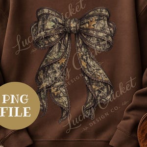 May include: A brown sweatshirt featuring a large bow design in a camouflage pattern. The bow has detailed shading and a decorative edge. The words "Lucky Cricket Design Co." are written on the sweatshirt, along with "PNG FILE" in a circle.