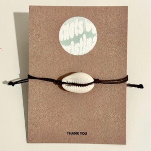 May include: A brown cord bracelet with a white cowrie shell charm. The bracelet is on a brown card with the text "Knots by Kristin" and "Thank You".