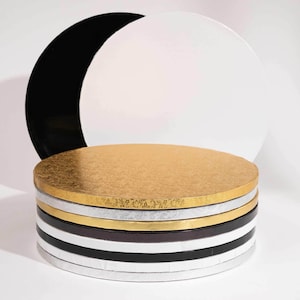 May include: A selection of round cake boards in gold, silver, black, white, and brown. Two large, solid-coloured boards in black and white are behind the stack. The boards are made of a sturdy material, suitable for supporting cakes.