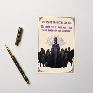 May include: A gothic birthday card with a purple and black design. The card features an illustration of a robed figure and figures with bat wings. The text on the card reads, "WE WISH TO INFORM YOU THAT YOUR BIRTHDAY HAS ARRIVED!" A black and gold pen is also in the image.