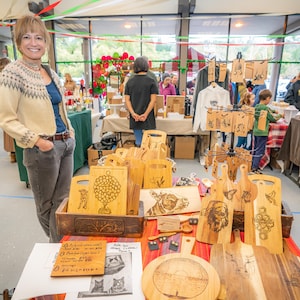 May include: A woman stands behind a table displaying wooden cutting boards with various designs, including a honeycomb, a butterfly, and a cat. The boards are arranged in a wooden crate and on a red and white tablecloth. A handwritten price list is visible on the table.