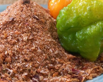 May include: A pile of brown spice blend with visible herbs and spices, next to a bright green and yellow pepper and a small orange pepper, all on a light blue plate. The spice blend appears coarse and textured.