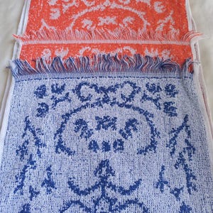 以下が含まれることがあります： フリンジ付きの縁が特徴の装飾用タオル2枚。1枚はオレンジ色に白い花柄、もう1枚は白地に青い花柄です。タオルは織り込まれた生地で作られています。