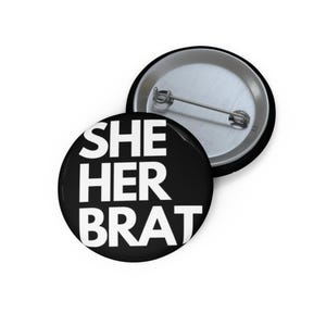 Könnte beinhalten: Zwei runde, schwarze Ansteckbuttons mit dem Text "SHE HER BRAT" in weißen, fetten Buchstaben. Ein Button liegt mit der Vorderseite nach oben, der andere mit der Rückseite nach unten, wobei die Nadel zu sehen ist. Die Buttons liegen auf einer weißen Oberfläche.