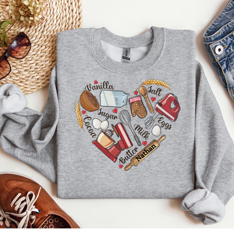 K&ouml;nnte beinhalten: Grauer Pullover mit einem herzf&ouml;rmigen Design, das verschiedene Backzutaten und -werkzeuge zeigt, darunter Vanille, Zucker, Kakao, Eier, Milch, Butter, Salz und einen Schneebesen. Der Text "Nathan" ist im Design enthalten.