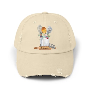 May include: A beige distressed baseball cap with a white background featuring a watercolor illustration of kitchen utensils, flowers, and the word "Grammy".