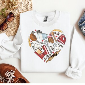 K&ouml;nnte beinhalten: Wei&szlig;er Pullover mit einem herzf&ouml;rmigen Design, das Backzutaten und -werkzeuge zeigt. Die Zutaten umfassen Vanille, Zucker, Kakao, Eier, Salz, Milch und Butter. Die Werkzeuge umfassen einen Schneebesen, einen Nudelholz, R&uuml;hrsch&uuml;sseln und Ofenhandschuhe. Der Text "Abby" steht am unteren Rand des Herzens.