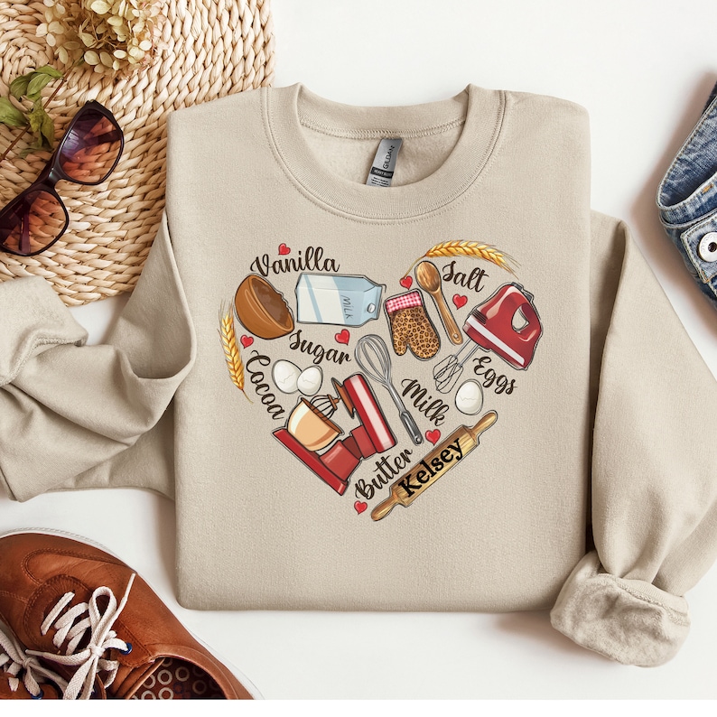 K&ouml;nnte beinhalten: Beigefarbener Pullover mit einem herzf&ouml;rmigen Design, das Backzutaten und -werkzeuge zeigt. Die Worte "Vanilla, Sugar, Cocoa, Butter, Milk, Salt, Eggs, Kelsey" sind um das Herz herum geschrieben.