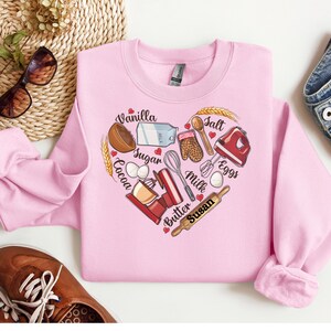 K&ouml;nnte beinhalten: Rosa Sweatshirt mit einem herzf&ouml;rmigen Design mit Backzutaten und -werkzeugen. Die Zutaten umfassen Vanille, Zucker, Kakao, Eier, Milch, Butter und Salz. Die Werkzeuge umfassen einen Schneebesen, einen Nudelholz, eine R&uuml;hrsch&uuml;ssel, einen Messbecher, einen Spatel und Ofenhandschuhe. Der Text "Susan" steht in der Mitte des Herzens.