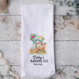 May include: White kitchen towel with a blue and white stand mixer with floral accents. The text "Gabby's Baking Co. Open daily" is printed below the mixer.