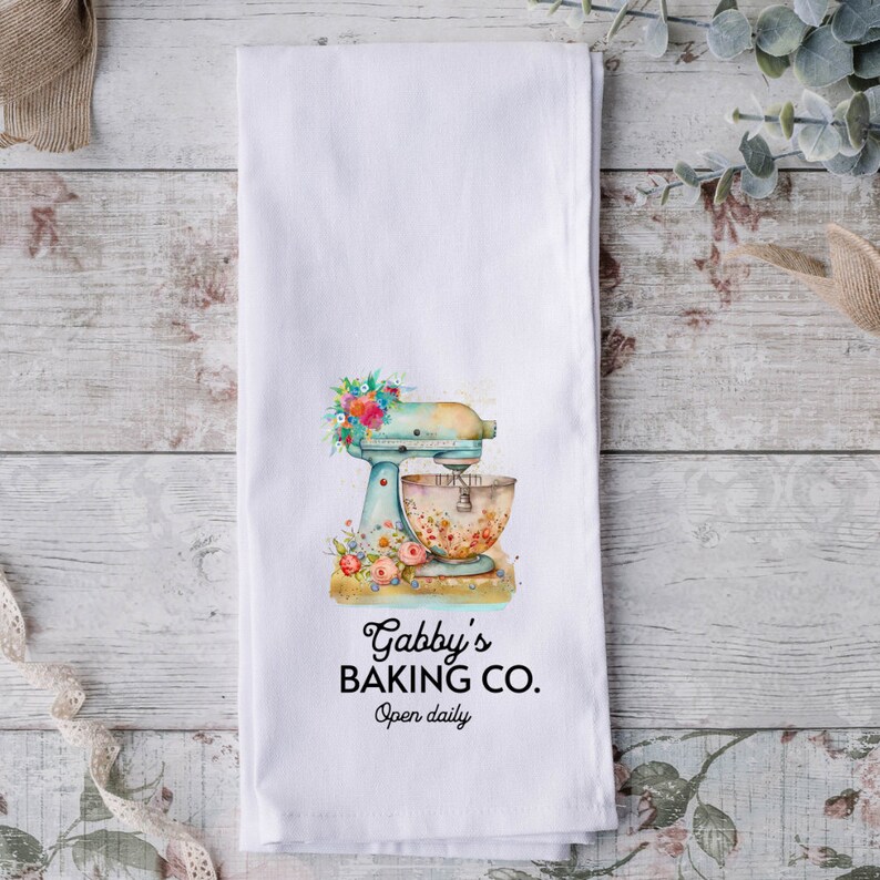 May include: White kitchen towel with a blue and white stand mixer with floral accents. The text "Gabby's Baking Co. Open daily" is printed below the mixer.