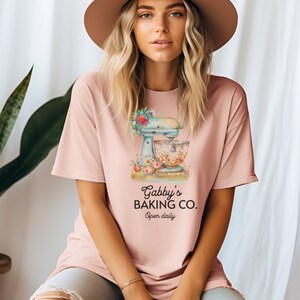May include: A pink t-shirt with a graphic of a vintage stand mixer with flowers and the text "Gabby's Baking Co. Open daily."