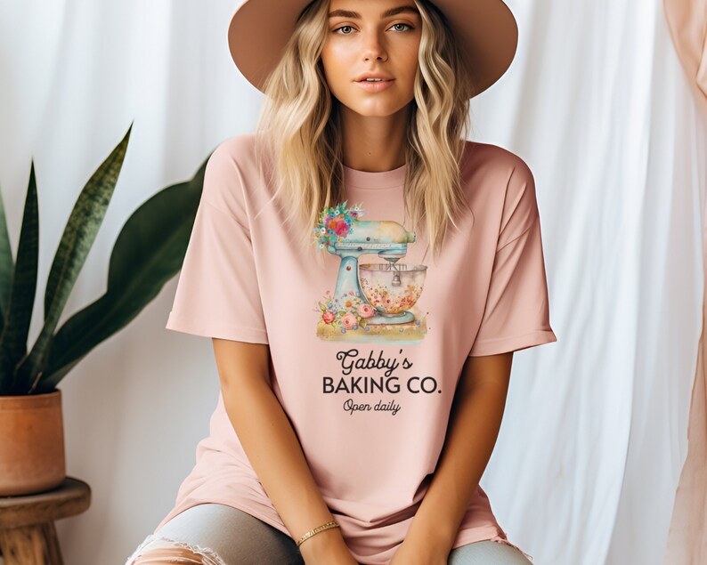 May include: A pink t-shirt with a graphic of a vintage stand mixer with flowers and the text "Gabby's Baking Co. Open daily."