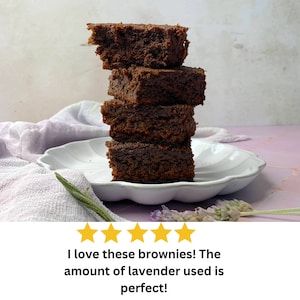 May include: A stack of five chocolate brownies on a white plate with a purple cloth in the background. The brownies are arranged in a tower, with the top brownie slightly broken. The image has a five-star rating and the text "I love these brownies! The amount of lavender used is perfect!"