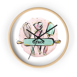 May include: A white round clock with a wooden frame. The clock face features a pink watercolor background with gold glitter and a green rolling pin with the name "Grace" on it. There are also images of a whisk, a pastry bag, and a spatula.