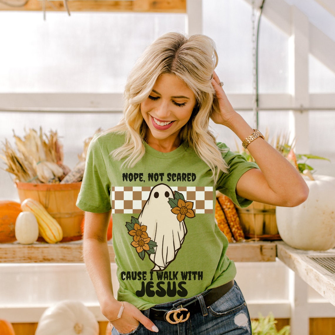May include: Green t-shirt with a white ghost and brown flowers. The text on the shirt reads 'Nope, not scared cause I walk with Jesus'.