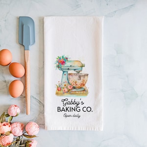 May include: White kitchen towel with a blue and white stand mixer with floral accents. The text "Gabby's Baking Co. Open daily" is printed on the towel.