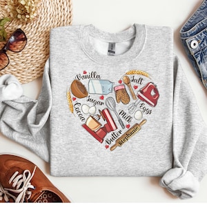 K&ouml;nnte beinhalten: Grauer Pullover mit einem herzf&ouml;rmigen Design mit Backzutaten und -werkzeugen. Die Zutaten umfassen Vanille, Zucker, Kakao, Eier, Milch, Butter und Salz. Die Werkzeuge umfassen einen Schneebesen, einen Nudelholz, eine Sch&uuml;ssel, einen Messbecher und ein Paar Ofenhandschuhe. Der Text "Stephanie" steht unter dem Herzen.