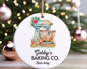 May include: A white ceramic ornament with a blue and white stand mixer with a floral design. The text "Gabby's Baking Co. Open daily" is printed on the ornament.