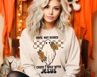 May include: Beige sweatshirt with a ghost graphic, floral accents, and the text "Nope, Not Scared Cause I Walk With Jesus." The ghost design is set against a brown and white checkered background. A casual, comfortable top.