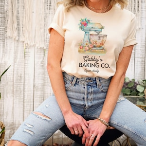 May include: A woman wearing a light beige t-shirt with a floral design and a blue stand mixer. The text on the shirt reads "Gabby's Baking Co. Open daily."