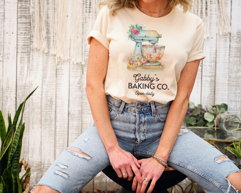 May include: A woman wearing a light beige t-shirt with a floral design and a blue stand mixer. The text on the shirt reads "Gabby's Baking Co. Open daily."