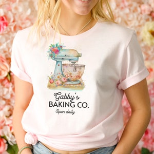 May include: A pink t-shirt with a blue and white stand mixer with floral accents. The text "Gabby's Baking Co. Open daily" is printed on the shirt.