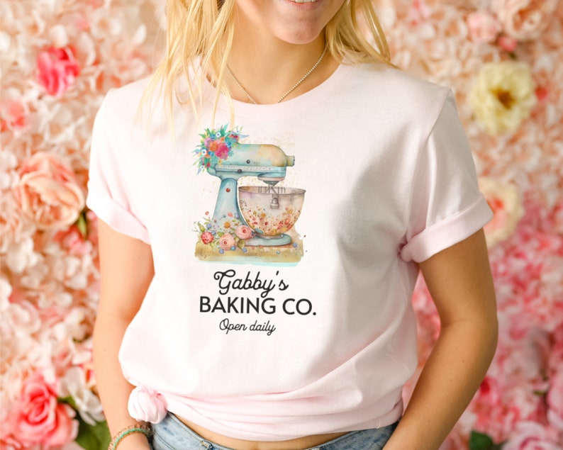 May include: A pink t-shirt with a blue and white stand mixer with floral accents. The text "Gabby's Baking Co. Open daily" is printed on the shirt.