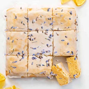 May include: Overhead shot of a square-cut lemon cake with a white glaze, decorated with lavender buds. Two slices are cut and separated from the rest. The cake sits on a white surface, with lemon slices in the corner.