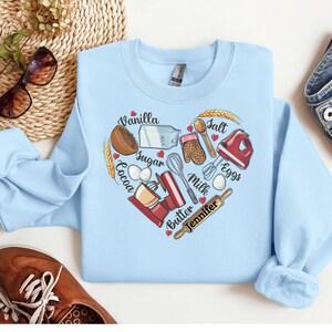 K&ouml;nnte beinhalten: Hellblauer Pullover mit einem herzf&ouml;rmigen Design, das Backzutaten und -werkzeuge zeigt. Die Zutaten umfassen Vanille, Zucker, Kakao, Eier, Milch, Butter und Salz. Die Werkzeuge umfassen einen Schneebesen, einen Nudelholz, eine R&uuml;hrsch&uuml;ssel, einen Holzl&ouml;ffel und Ofenhandschuhe. Der Text "Jennifer" steht unter dem Herzen.