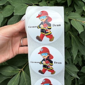 Pode incluir: Um rolo de adesivos redondos brancos com um desenho animado de um bombeiro segurando uma mangueira de inc&ecirc;ndio. Os adesivos t&ecirc;m as palavras "Bombeiro" e "Dean" impressas neles.