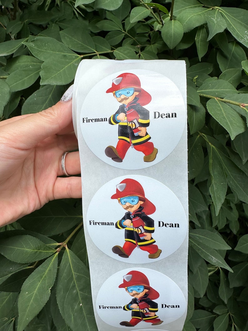 Pode incluir: Um rolo de adesivos redondos brancos com um desenho animado de um bombeiro segurando uma mangueira de inc&ecirc;ndio. Os adesivos t&ecirc;m as palavras "Bombeiro" e "Dean" impressas neles.
