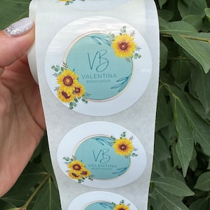 Pode incluir: Rolo de adesivos redondos com fundo azul claro, um c&iacute;rculo dourado e o texto "VB Valentina Beachwear" com um design de girassol.