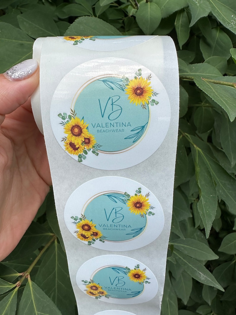 Pode incluir: Rolo de adesivos redondos com fundo azul claro, um c&iacute;rculo dourado e o texto "VB Valentina Beachwear" com um design de girassol.