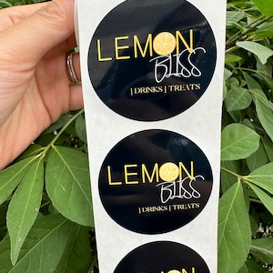 Pode incluir: Um rolo de adesivos redondos pretos e brancos com o texto "LEMON BLISS | DRINKS | TREATS" e um gr&aacute;fico de fatia de lim&atilde;o amarelo.