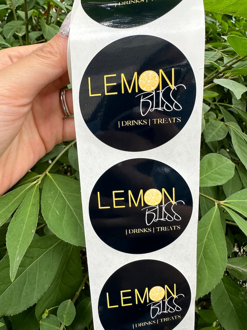Pode incluir: Um rolo de adesivos redondos pretos e brancos com o texto "LEMON BLISS | DRINKS | TREATS" e um gr&aacute;fico de fatia de lim&atilde;o amarelo.