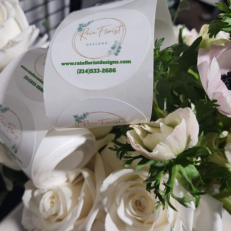 Pode incluir: Flores brancas e rosadas com folhas verdes. Um adesivo redondo com o texto "Rain Florist Designs" e o endere&ccedil;o do site "www.rainfloristdesigns.com" e o n&uacute;mero de telefone (214) 533-2686.