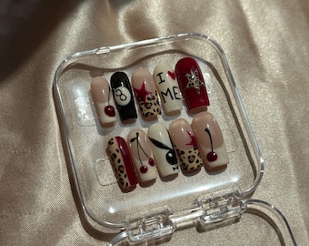 May include: A set of press-on nails in a clear plastic case. The nails feature various designs, including cherries, leopard print, the number 8, the text "I ❤️ ME", and a Playboy bunny logo. The nails are in various colors, including red, black, and nude.