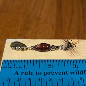 Może przedstawiać: Brązowo-złota przynęta wędkarska z pi&oacute;rkowatym ogonem, o długości około 2,5 cm. Przynęta leży na drewnianej powierzchni z linijką do skalowania.