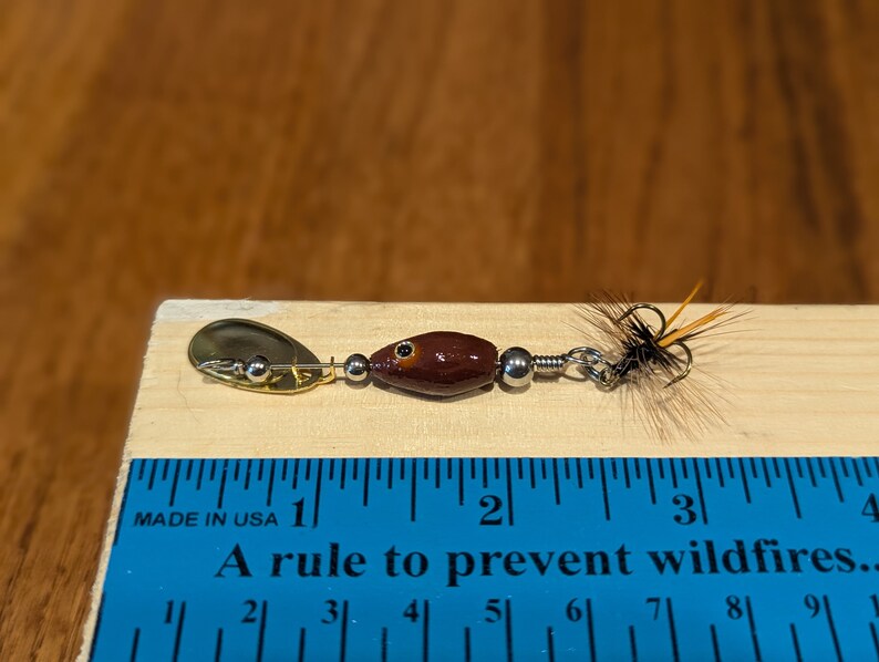 Może przedstawiać: Brązowo-złota przynęta wędkarska z pi&oacute;rkowatym ogonem, o długości około 2,5 cm. Przynęta leży na drewnianej powierzchni z linijką do skalowania.