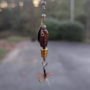 May include: Close-up of a fishing lure with a brown body, gold accents, and a feathered hook. The lure is suspended by a thin wire against a blurred outdoor background. The lure is designed for catching fish.
