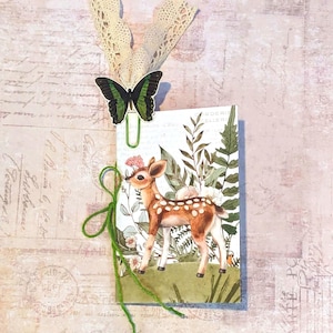 May include: A decorative tag featuring a fawn illustration with a green and white floral background. A green butterfly paperclip and a cream-colored lace ribbon are attached to the tag. A green string is tied around the tag.