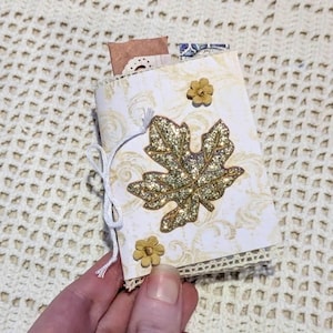 May include: A small handmade booklet with a cream-colored cover featuring a faint gold swirl pattern. The front is decorated with a large gold glitter maple leaf, two small beige flower embellishments, and a white string tied in a bow.