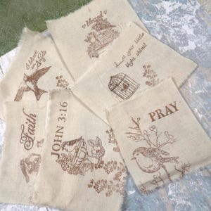 May include: A collection of beige burlap fabric pieces with brown ink illustrations and text. Designs include birds, floral elements, a birdcage, and phrases like "Faith", "Pray", and "Blessed". The edges are frayed.